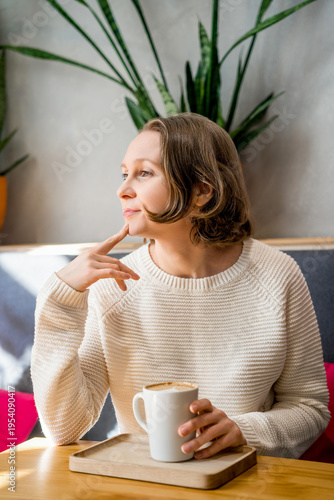 A thoughtful woman in a white knitted sweater sits in a cafe, holding a coffee cup. Peaceful atmosphere in a bright, modern interior.