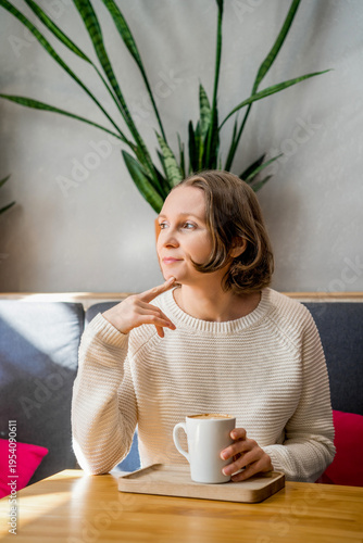 A thoughtful woman in a white knitted sweater sits in a cafe, holding a coffee cup. Peaceful atmosphere in a bright, modern interior.