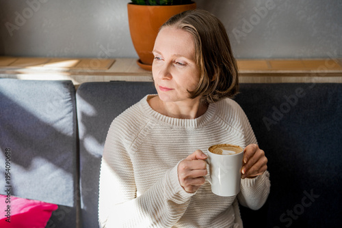 A thoughtful woman in a white knitted sweater sits in a cafe, holding a coffee cup. Peaceful atmosphere in a bright, modern interior.