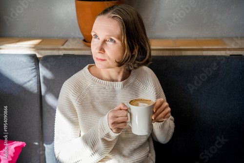 A thoughtful woman in a white knitted sweater sits in a cafe, holding a coffee cup. Peaceful atmosphere in a bright, modern interior.