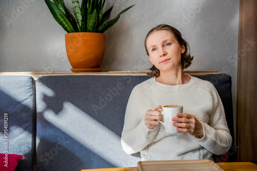 A thoughtful woman in a white knitted sweater sits in a cafe, holding a coffee cup. Peaceful atmosphere in a bright, modern interior.