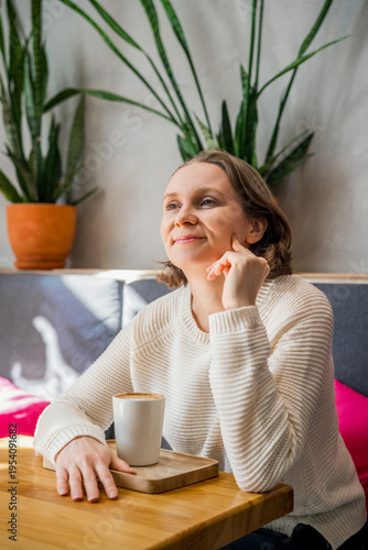 A thoughtful woman in a white knitted sweater sits in a cafe, holding a coffee cup. Peaceful atmosphere in a bright, modern interior.