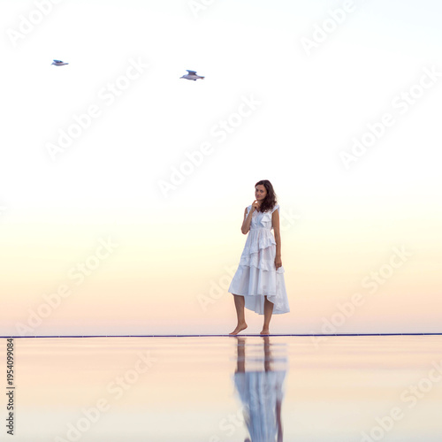 Картината върху платно A woman in a white dress stands on the mirror-like surface of the water or by an infinity pool at sunset