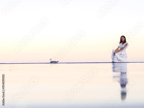 Fotografie A woman in a white dress stands on the mirror-like surface of the water or by an infinity pool at sunset
