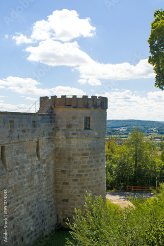 Sunlit stone tower rises above the old fortress wall in Altenburg, Germany. Soft clouds drift across a bright blue sky over rolling green hills beyond. Quiet paths and trees frame the historic scene.