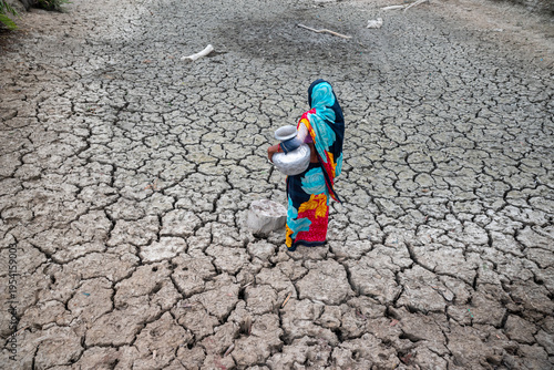 Satkhira, Bangladesh - 25 April 2025: View of a lone woman carrying water vessels across a parched, cracked earth, her vibrant clothing a stark contrast to the arid landscape.
