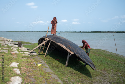 Satkhira, Bangladesh - 26 April 2025: View of a weathered, upturned boat resting on the grassy riverbank, figures tending to it under a vast sky.