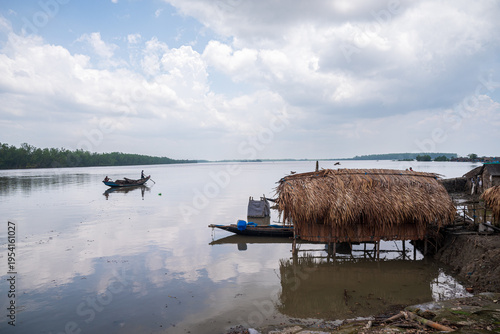 Khulna, Bangladesh - 12 July 2025: View of a stilt hut's thatched roof reflecting on the still water, as a lone boatman navigates the waterway.