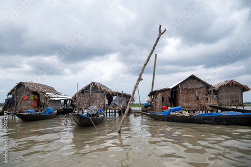Khulna, Bangladesh - 13 July 2025: View of stilted homes reflecting on the water's surface, boats moored, under a heavy, overcast sky, portraying the resilience of coastal life.
