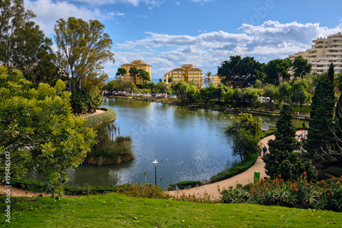 Benalmadena, Spain - 24 December 2025: View of Parque de la Paloma's tranquil lake reflecting the clear sky, embraced by vibrant green foliage and distant buildings.
