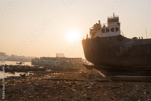 Keraniganj, Bangladesh - 11 February 2023: View of hulking ship beside smaller boats along the shoreline, reflecting the golden sunlight in a poignant scene.