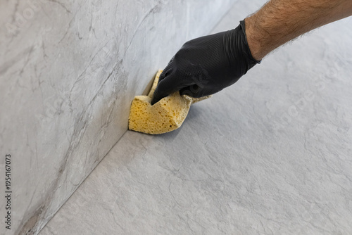 Obraz na plátně Worker in a black nitrile glove cleaning grout residue from gray porcelain tiles with a yellow sponge during kitchen renovation