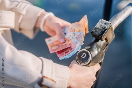 Female holding banknotes while preparing to refuel a vehicle at a gas station, fuel nozzle in hand, with blurred background of the station and surroundings