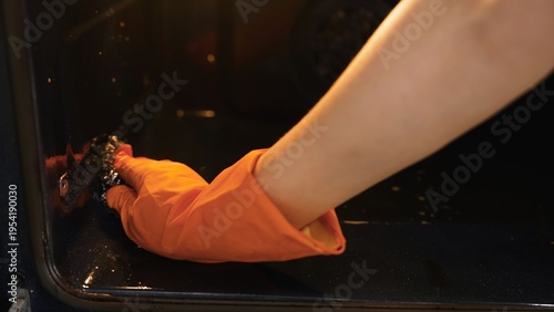 Bild auf Leinwand Close up of a person in an orange rubber glove scrubbing the inside of a dirty kitchen oven with a scouring pad