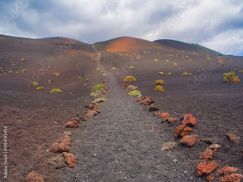 Volcanic ash trail leading up a dramatic slope - A hiking trail lined with red volcanic rocks ascends a dramatic ash slope with sparse vegetation under a cloudy sky on the Ruta de los Volcanes.