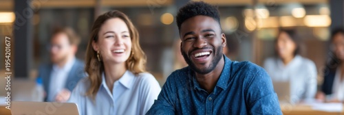 Two young professionals smiling and enjoying a lively conversation in a modern office setting with blurred coworkers in the background.