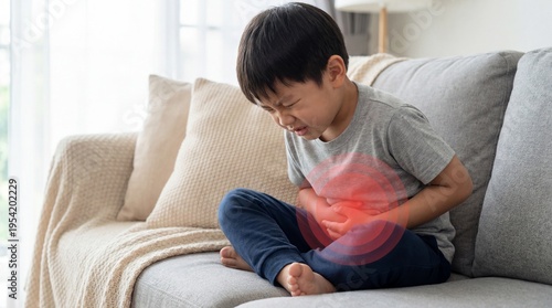 Asian boy sitting on sofa suffering from stomach ache with red pained area effect on abdomen