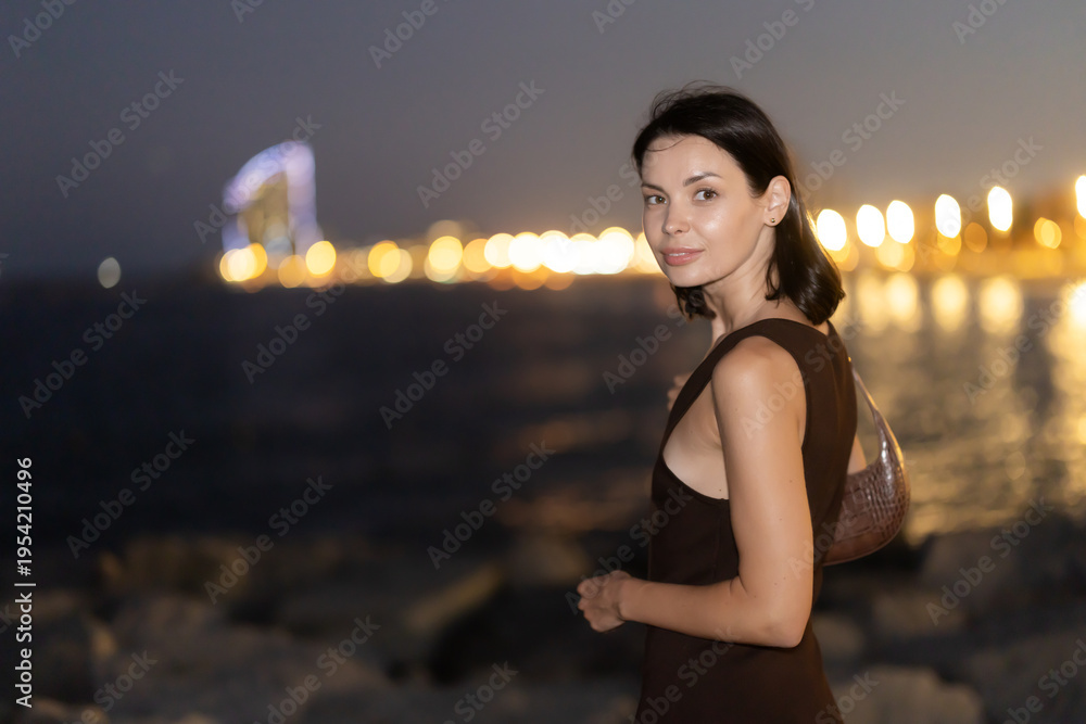 Fototapeta premium Female tourist is exploring citys attractions, brightly lit by electricity. Woman at waters edge, hotel skyscraper Arts in background. Night fell on city..