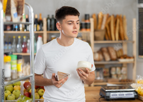 Konstfotografi Attentive young guy is choosing different types of cheese in grocery store