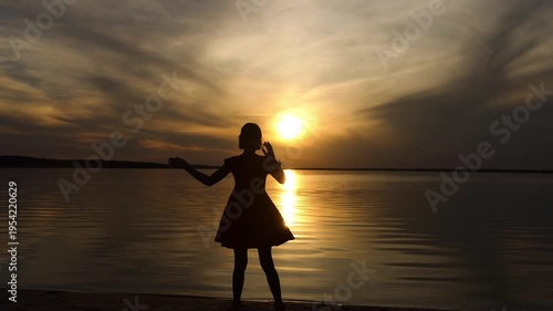 Silhouette of a little dancing girl against the background of the setting sun and a large lake. Beautiful sunset.