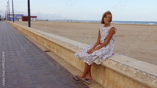 Little girl sitting on a stone curb raised sandy beach near the sea in a white dress.