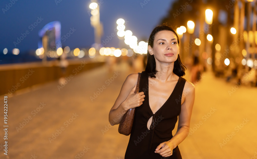 Fototapeta premium At night, slender female brunette walks in vicinity of beach in Barceloneta, spends time in motion, walks and enjoys sunny warm day. Carefree, lively nightlife, promenade..