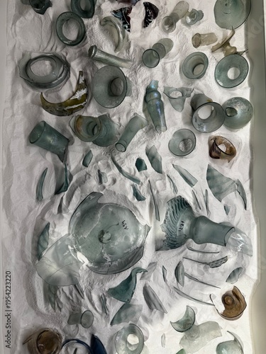 A curated collection of ancient glass artifacts and broken fragments carefully displayed on a textured white surface, resembling an archaeological excavation. The image highlights historical craftsman