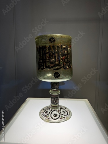 An elegant antique goblet featuring intricate traditional patterns and handcrafted details, displayed on a modern illuminated pedestal. The contrast between historical craftsmanship and contemporary p