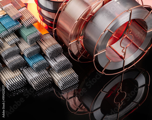 Industrial welding consumables with electrodes and wire coils on reflection. High angle view of welding rods in boxes and copper wire spools.