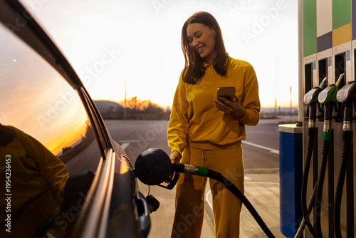 Konstfotografi Happy woman driver filling her car with fuel at the gas station and using cellph