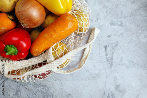 Colorful assortment of fresh farm produce including bell peppers onion and carrots in an ecobag on a grey background