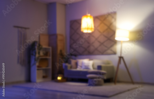 Blurred view of modern living room with grey sofa, glowing lamps and stylish carpet hanging on wall at evening