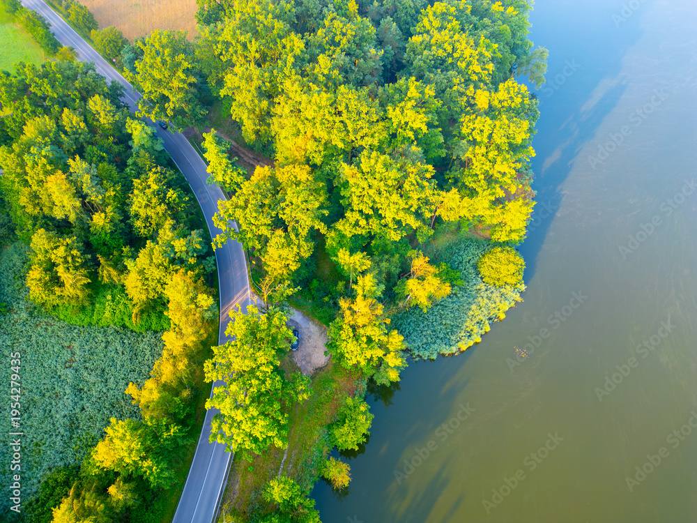 Fototapeta premium A winding road runs alongside a body of water. Trees surround the area with a mix of green foliage. The scene is captured from an aerial view during daylight.