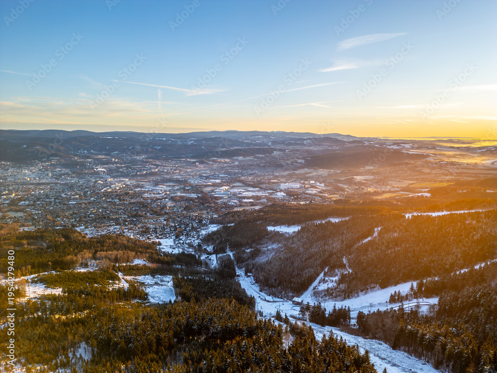 Fototapeta premium The scene shows a winter morning landscape in Liberec from a drone's view. Snow-covered hills and valleys stretch across the area with a warm light from the rising sun.