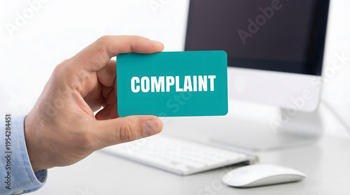 A human hand in a blue shirt holds a bright turquoise card with the word COMPLAINT clearly visible in a brightly lit professional office setting featu