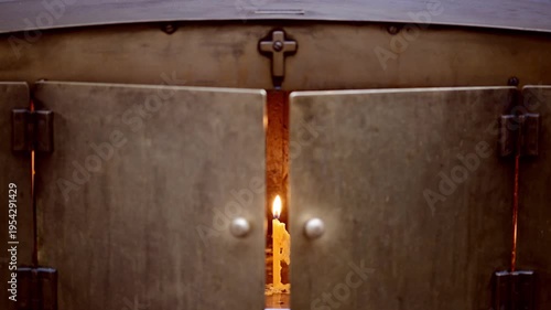 A candle burns softly, casting warm light through the gap of heavy metal doors