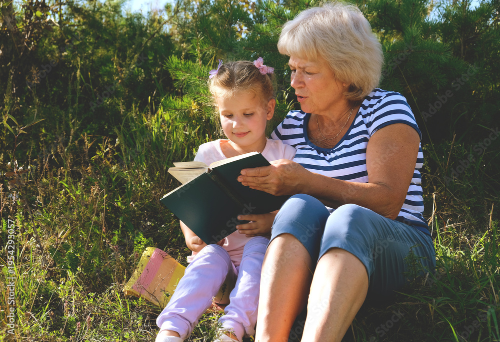 Fototapeta premium Happy grandmother and granddaughter reading book of stories or novel at leisure while sitting in the garden, backyard. Grandmother delighting in storytelling, engaged in reading a book