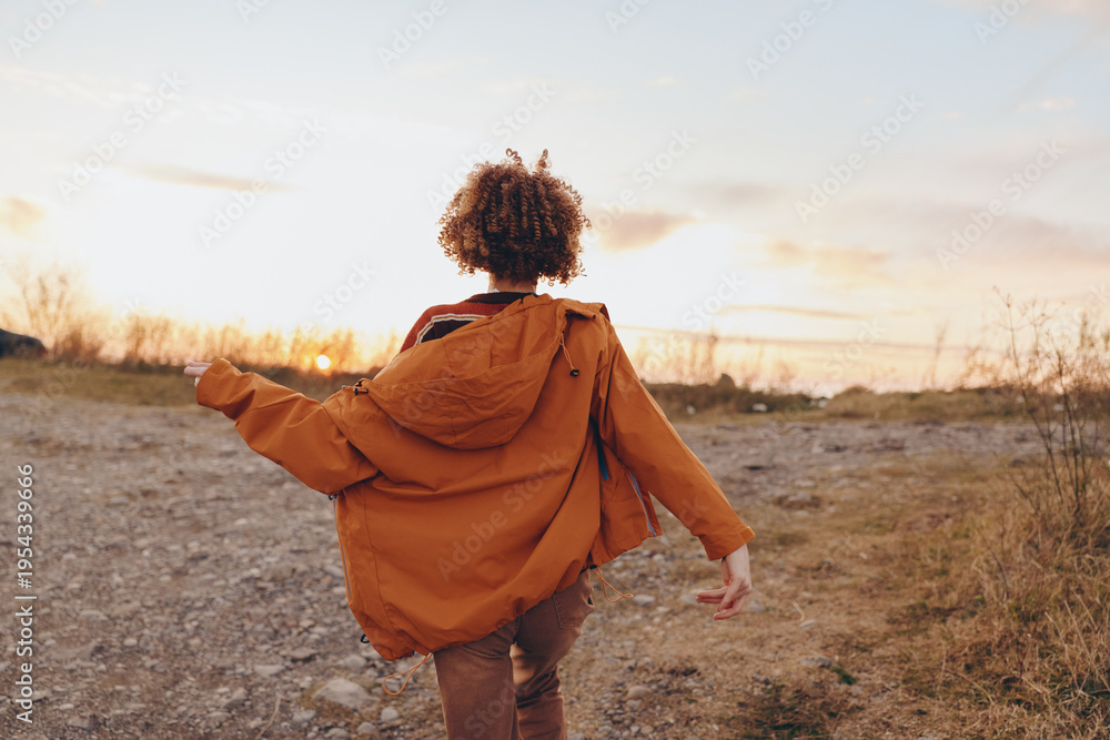 Fototapeta premium A woman wearing an orange jacket walks on a rocky path during sunset. The woman enjoys nature and freedom in the open landscape with a clear sky and warm light.