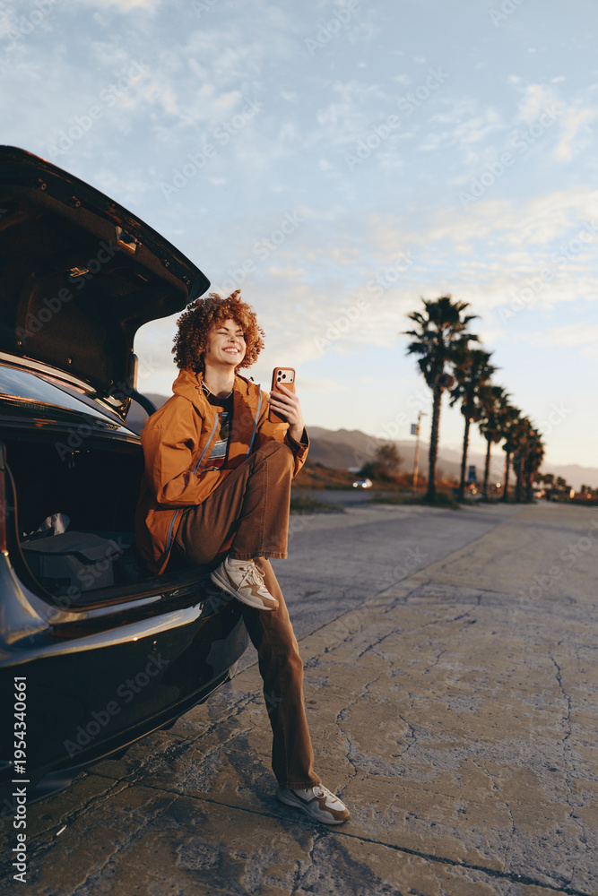 Naklejka premium Young woman with curly hair sitting in car trunk smiling, holding smartphone, wearing casual clothes, outdoor lifestyle, palm trees in background, warm sunlight, relaxed mood.