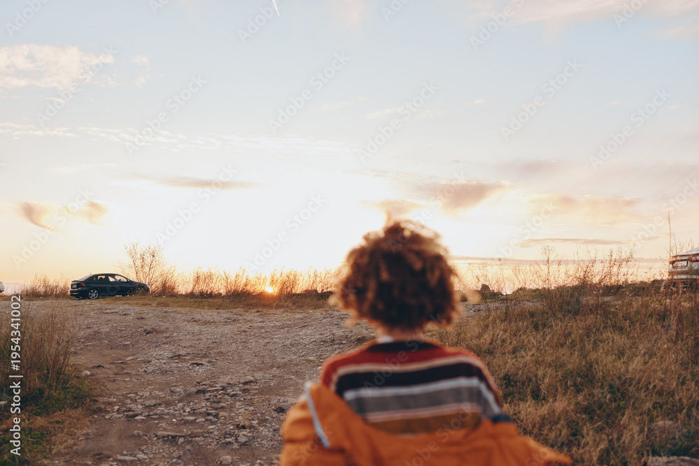 Fototapeta premium Woman wearing rainbow sweater stands outdoors near car at sunset, holding smartphone, enjoying lifestyle moment with warm light and natural landscape in background.