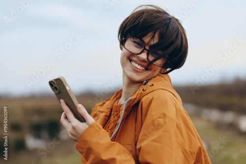 Smiling woman lifestyle wearing glasses and orange jacket holds smartphone outdoors in natural environment. Happy young female enjoys technology and casual fashion concept.