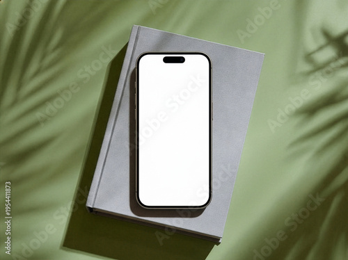 Minimalist flat lay of a blank smartphone mockup on a grey book with palm leaf shadows on a green background