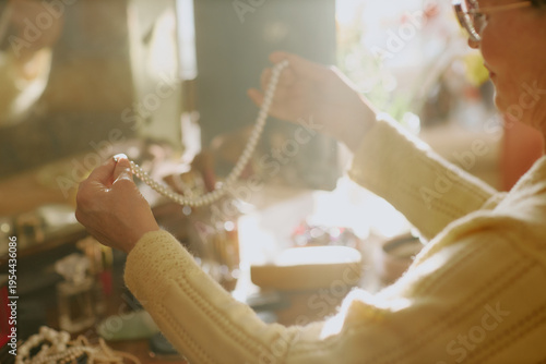 Senior Caucasian woman holding pearl necklace with both hands, examining jewelry at dressing table, sunlight illuminating hands and accessories, only upper body and hands visible