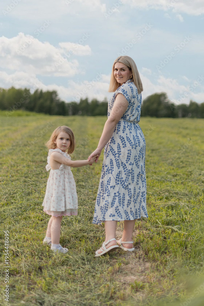 Fototapeta premium A young Caucasian woman and a small girl hold hands in a green field. The woman has long blonde hair and wears a patterned dress. The girl has short blonde hair and a white dress.
