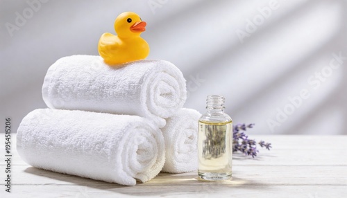 Stack of white towels with a yellow rubber duck on top, bottle of oil, and lavender on a white wooden surface in a bright bathroom setting