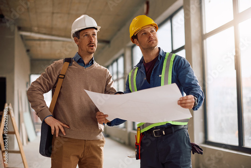 Project manager and worker cooperating while going through rebuilding plans at construction site.