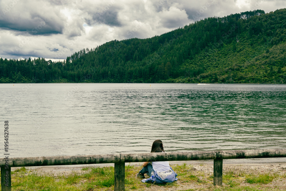 Fototapeta premium Woman Sitting Lakeside Near Forested Mountain