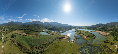 Aerial panoramic view of artificial fish ponds and wetlands surrounded by green hills and mountains in Santa Catarina, Brazil under a clear blue sky.
