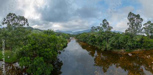 Aerial panoramic view of a calm river flowing through dense green forest with mountains in the background under a dramatic cloudy sky in Santa Catarina, Brazil.