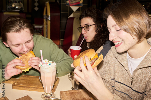 Canvas-taulu Three gen Z friends eating burgers and drinking milkshake at restaurant table, s